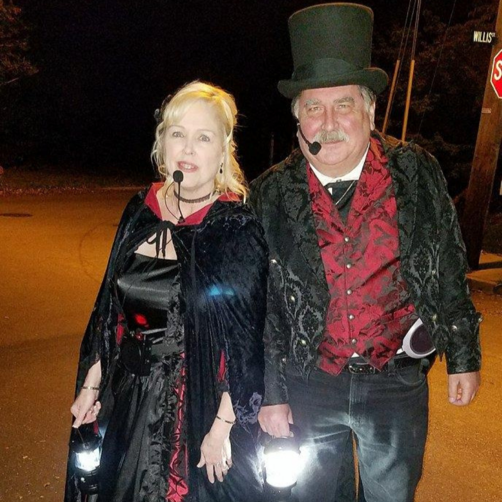 Two adults in Victorian-style costumes and capes stand outdoors at night during HALLOWEEN IN ACWORTH, each holding a lantern and wearing headsets. The man sports a top hat, with a stop sign visible in the background.