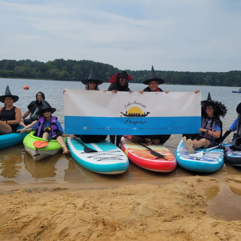 Eight people in witch hats sit on paddleboards at a lakeshore, holding a “Lake Norman Dragons” banner. Trees and water are visible in the background, capturing the festive spirit of Halloween in Acworth.