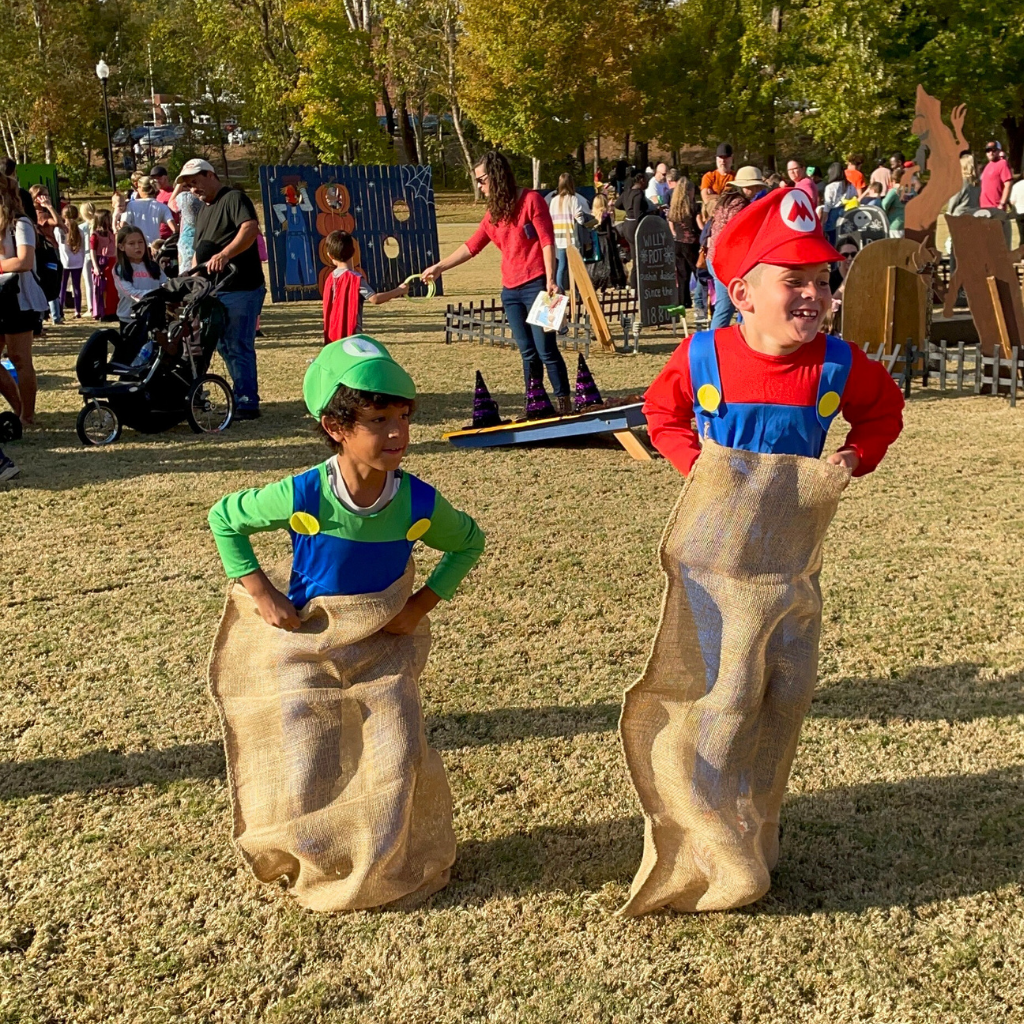 Two children dressed as Luigi and Mario join a sack race on the grass during HALLOWEEN IN ACWORTH, with festive people and decorations filling the outdoor event in the background.