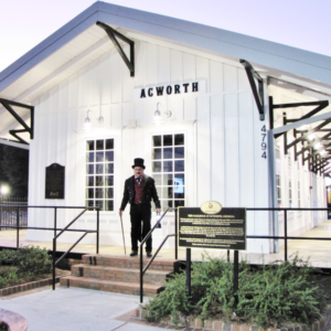 A man in a top hat and suit stands on the steps of a white building labeled “Acworth,” next to a historical marker and railings, capturing the mysterious spirit of Halloween in Acworth.