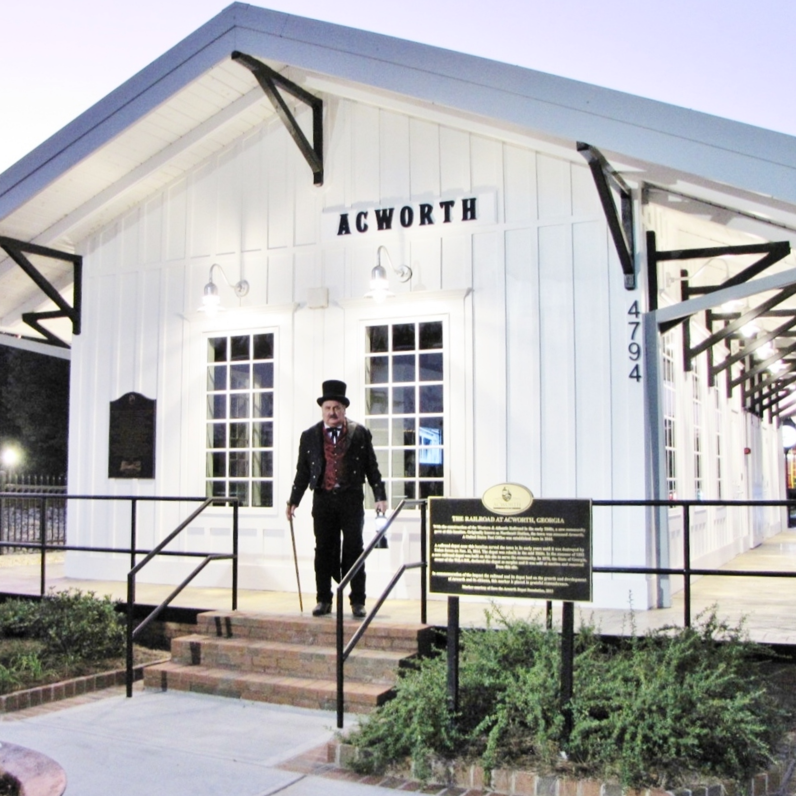 VisitAcworth Events SQUARE (14) A man in a top hat and suit stands on the steps of a white building labeled “Acworth,” next to a historical marker and railings, capturing the mysterious spirit of Halloween in Acworth.