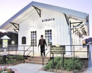 A man in a period costume stands on the steps of the historic white Acworth train depot, with plaques and signage visible outside the building, bringing to mind the festive spirit of Halloween in Acworth.