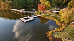 Aerial view of a lake with a wooden dock, bridge, and people walking on pathways and Trails in Acworth, all surrounded by trees with vibrant autumn foliage.