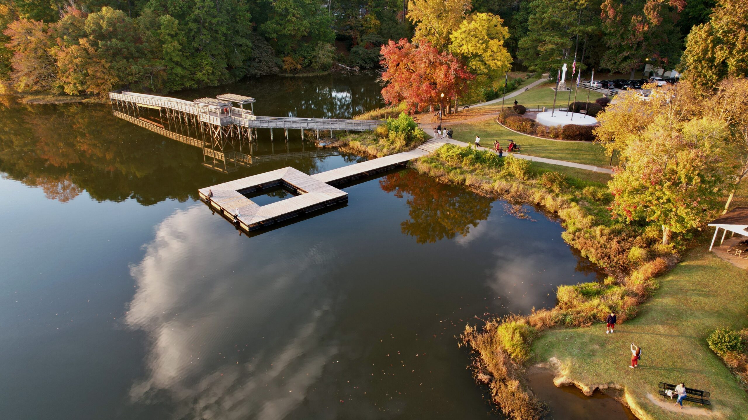 default Aerial view of a lake with a wooden dock, bridge, and people walking on pathways and Trails in Acworth, all surrounded by trees with vibrant autumn foliage.