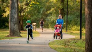 A woman pushes a stroller with a baby on a park path along the trails in Acworth, while a child rides a scooter ahead and another person jogs in the distance.
