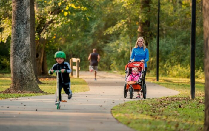 A woman pushes a stroller with a baby on a park path along the trails in Acworth, while a child rides a scooter ahead and another person jogs in the distance.