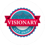 Circular badge with a city skyline in the background, featuring the text "VISIONARY" on a red banner and "CITY AWARD" below in a blue section.