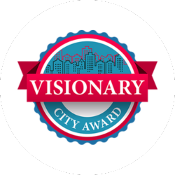 Circular badge with a city skyline in the background, featuring the text "VISIONARY" on a red banner and "CITY AWARD" below in a blue section.