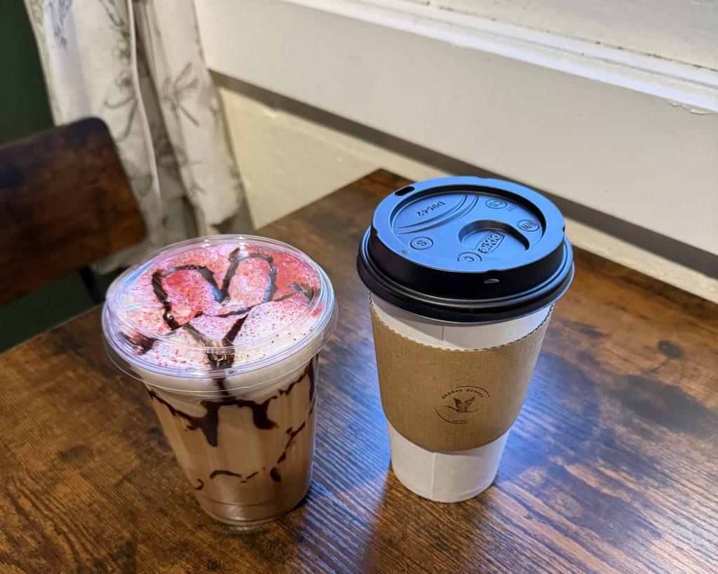 A plastic cup of iced chocolate drink with strawberry slices and chocolate drizzle next to a paper cup of hot coffee on a wooden table.