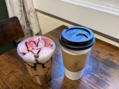 A plastic cup of iced chocolate drink with strawberry slices and chocolate drizzle next to a paper cup of hot coffee on a wooden table.