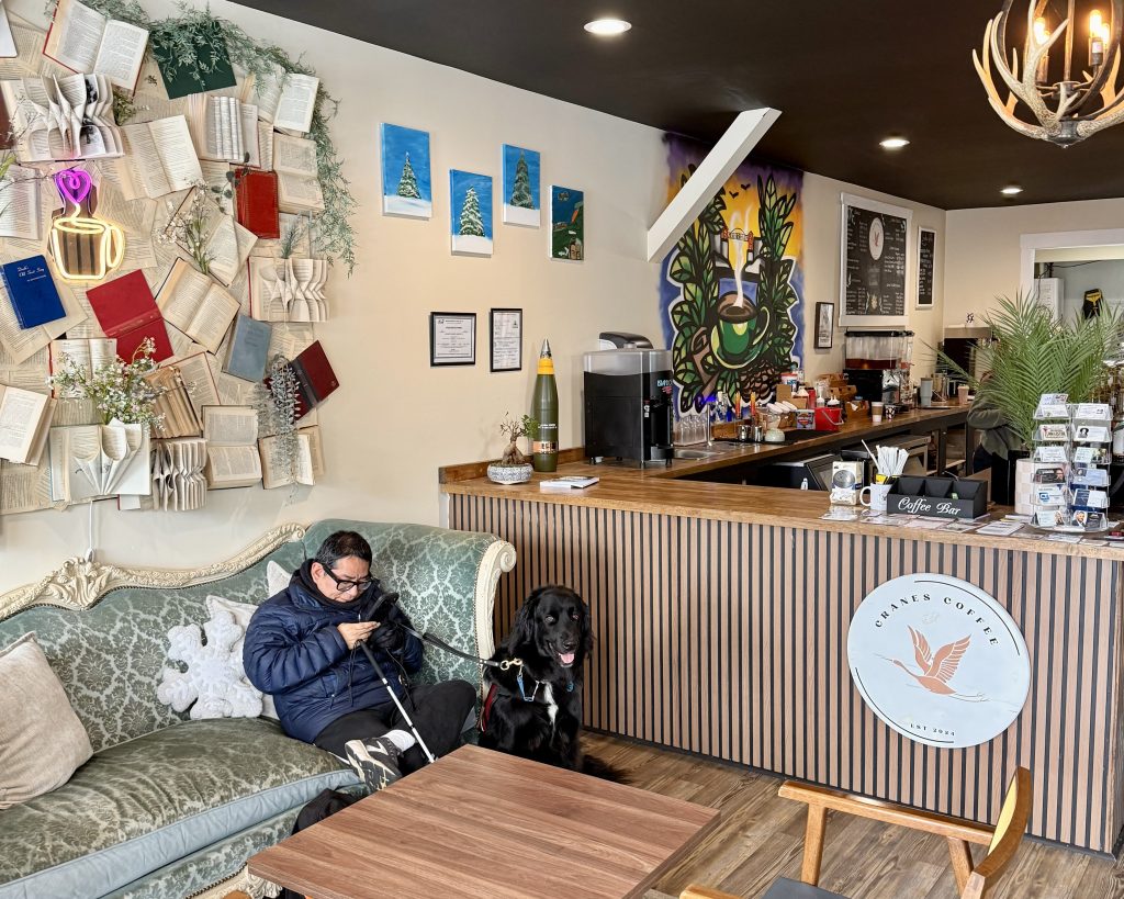 A person sits on a sofa using a phone next to a black dog in a cozy coffee shop, with books on the wall and art above the counter.