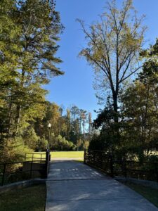 A paved path, part of the popular trails in Acworth, leads over a small bridge and into a grassy area, surrounded by trees with some autumn foliage under a clear blue sky.