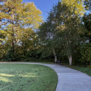 A curved concrete pathway, part of the scenic Trails in Acworth, winds through a park with green grass, tall trees, and a bench by a lamppost beneath a clear blue sky.