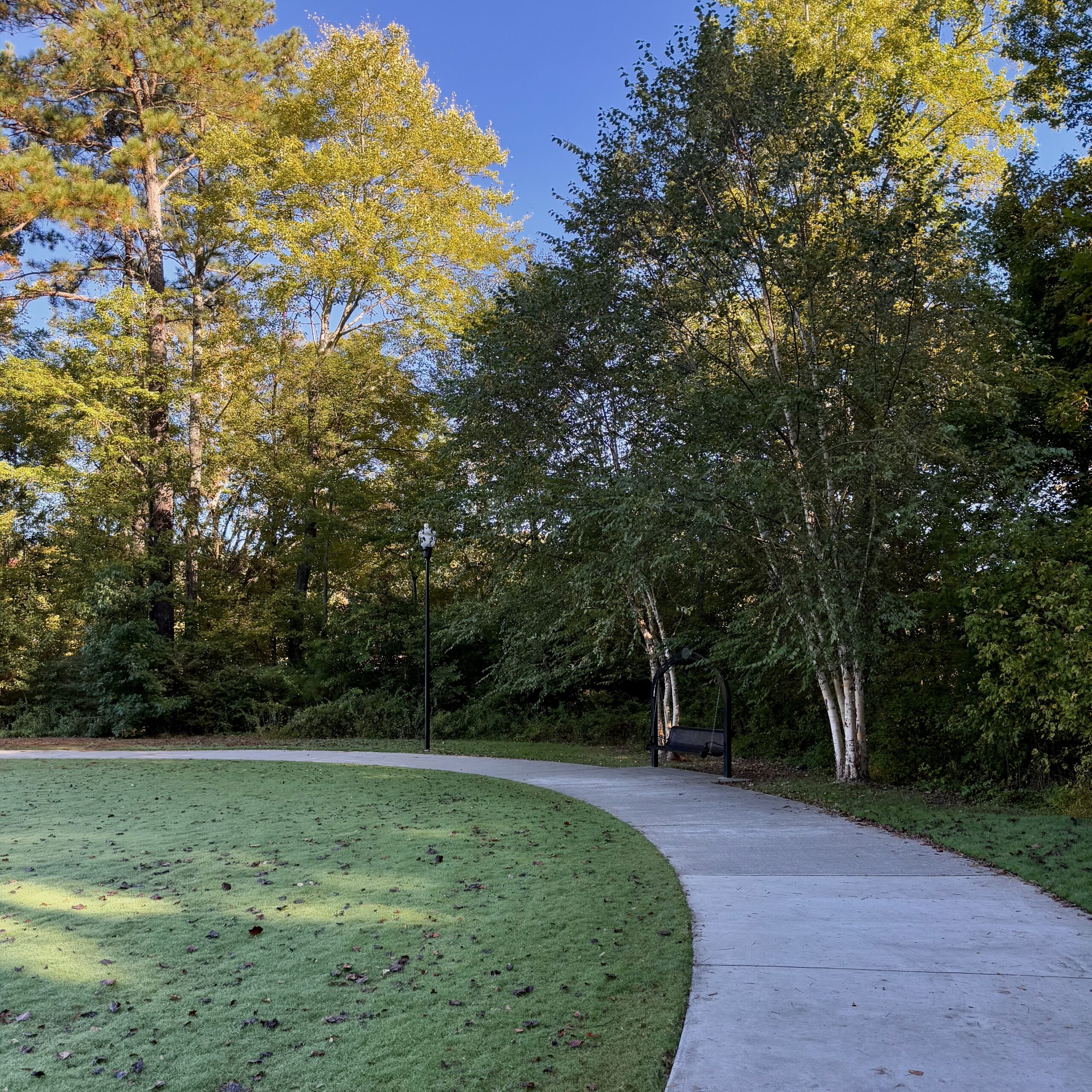 Photo Oct 15 2025, 10 01 39 AM A curved concrete pathway, part of the scenic Trails in Acworth, winds through a park with green grass, tall trees, and a bench by a lamppost beneath a clear blue sky.