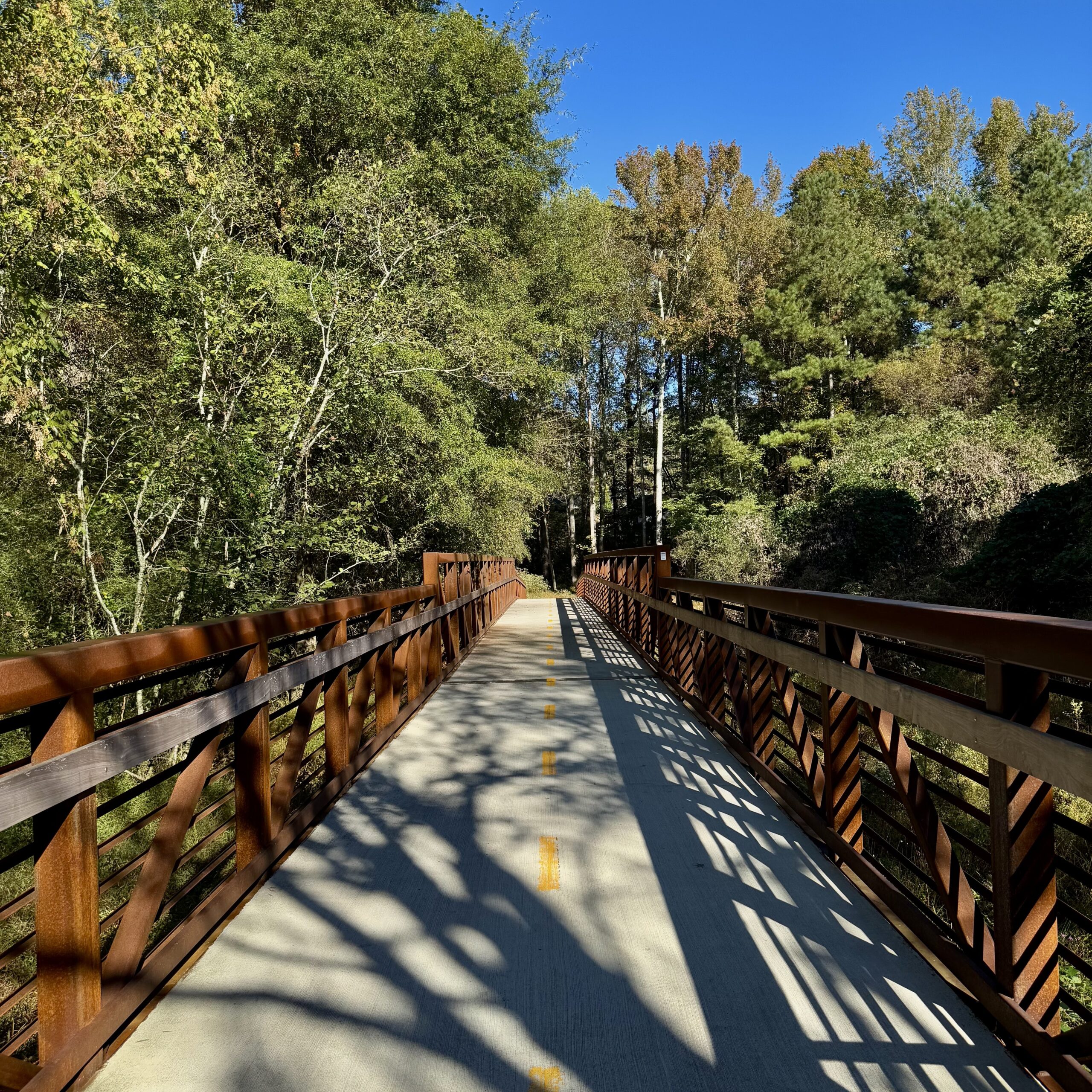 Photo Oct 15 2025, 10 23 28 AM A metal pedestrian bridge with a concrete path and yellow dividing line spans through a wooded area under a clear blue sky, inviting walkers to explore the scenic Trails in Acworth.
