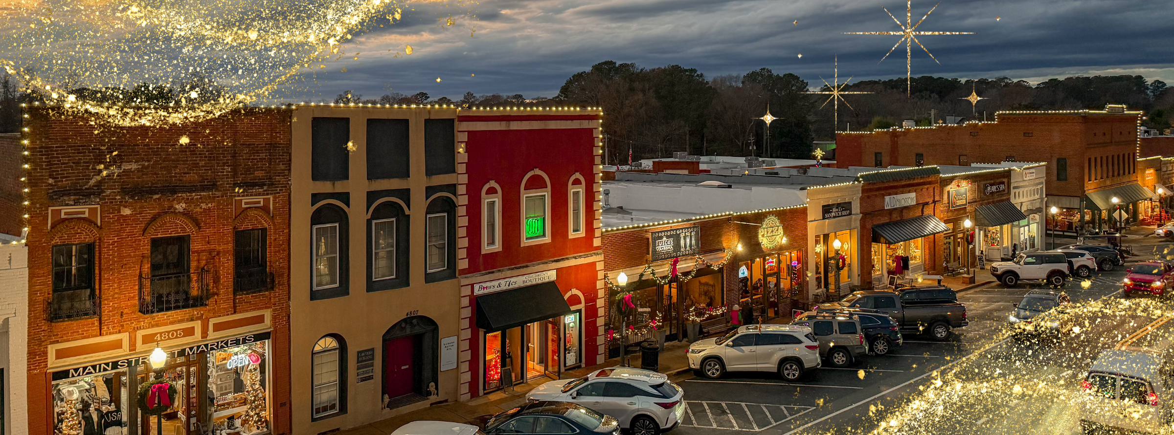 A small-town main street at dusk with holiday lights and decorations, storefronts, parked cars, and festive golden sparkles overlaying the image.