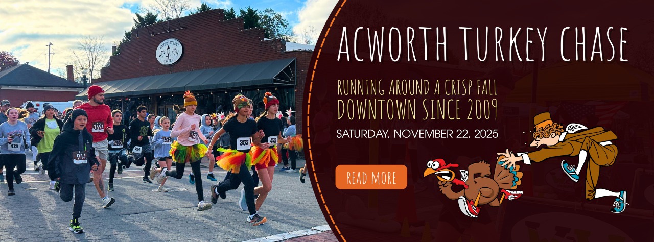 A group of people in costumes and running gear start a race in a downtown area under a clear sky; a banner promotes the Acworth Turkey Chase event.