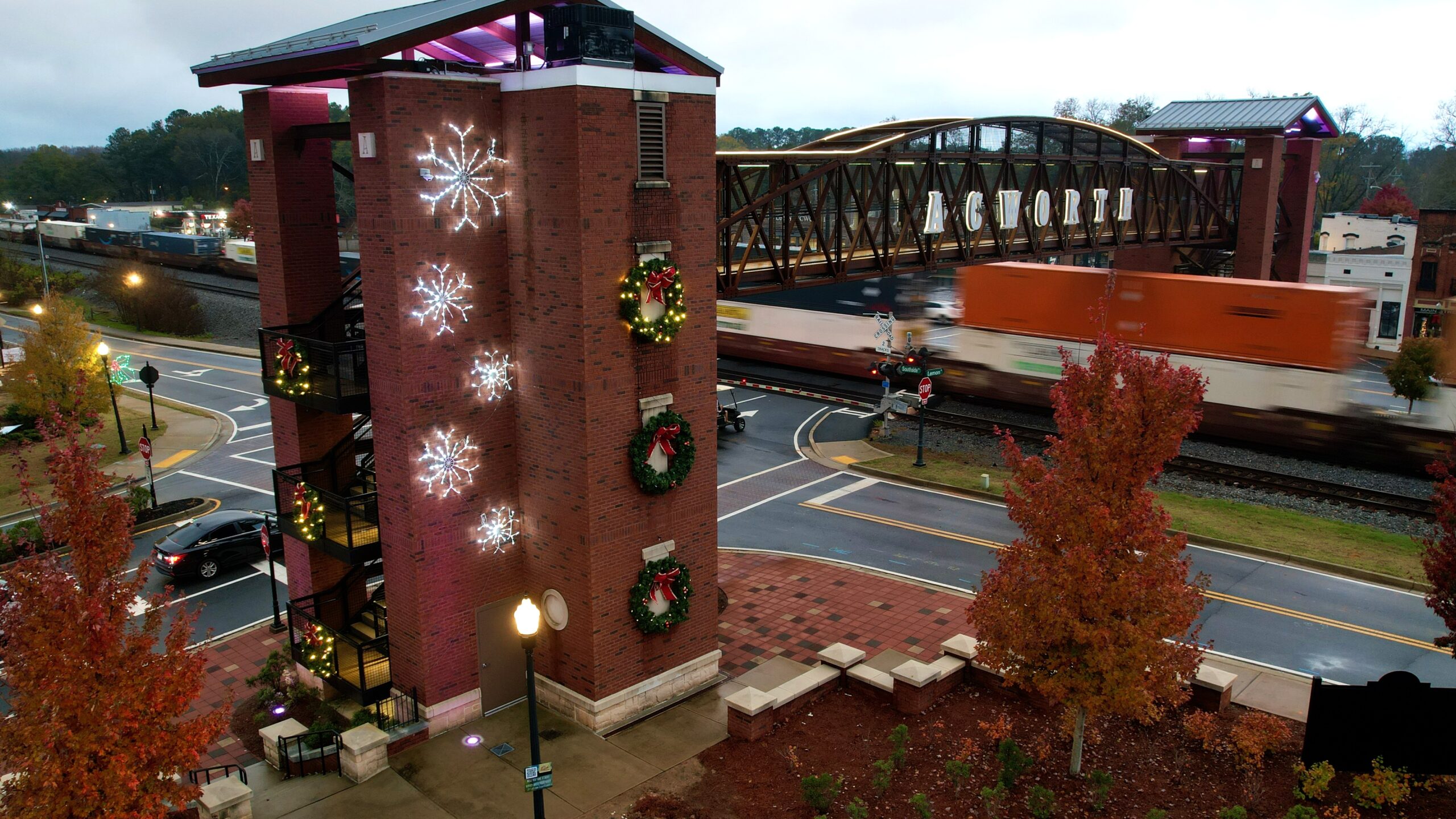 default A brick building adorned with wreaths and snowflake lights stands beside a railroad crossing, where a moving freight train passes under a large bridge labeled "ACWORTH." Your guide to Acworth gifts begins in this festive setting.