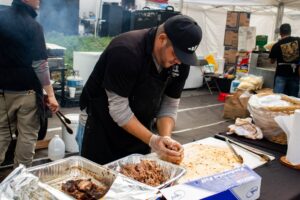 A man wearing gloves and a cap shreds cooked meat at an outdoor food stall, with trays of meat and other supplies on the table around him.