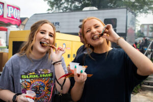 Two women are standing at an outdoor event, smiling and eating ribs, each holding a cup of dipping sauce. Food stands and a crowd are visible in the background.