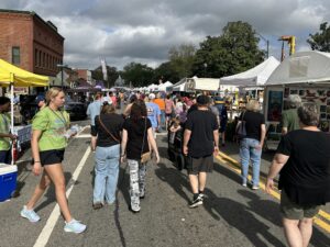 People walk through a street market with white tents and vendor booths on both sides under a partly cloudy sky.