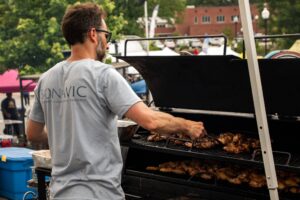A man wearing sunglasses grills chicken on an outdoor barbecue at a market or festival, surrounded by tents and trees.