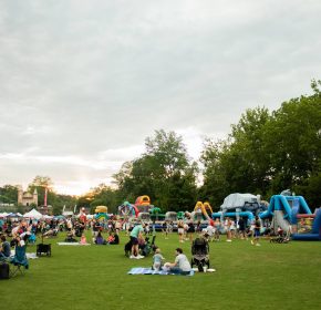 People gather on a grassy field with blankets and chairs at an outdoor event featuring tents and inflatable obstacle courses under a cloudy sky.