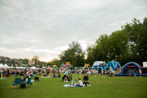 People gather on a grassy field with blankets and chairs at an outdoor event featuring tents and inflatable obstacle courses under a cloudy sky.