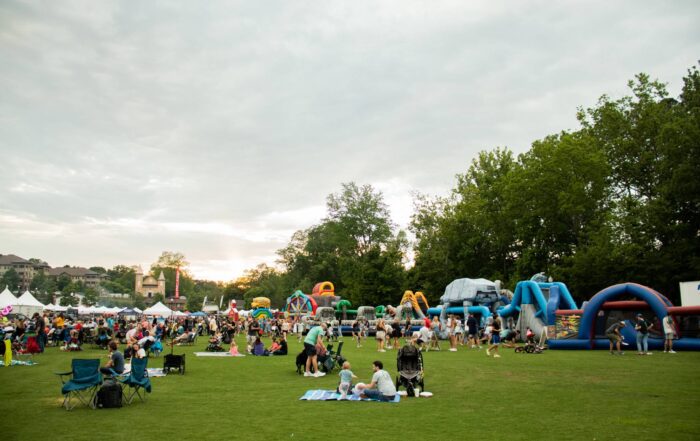 People gather on a grassy field with blankets and chairs at an outdoor event featuring tents and inflatable obstacle courses under a cloudy sky.