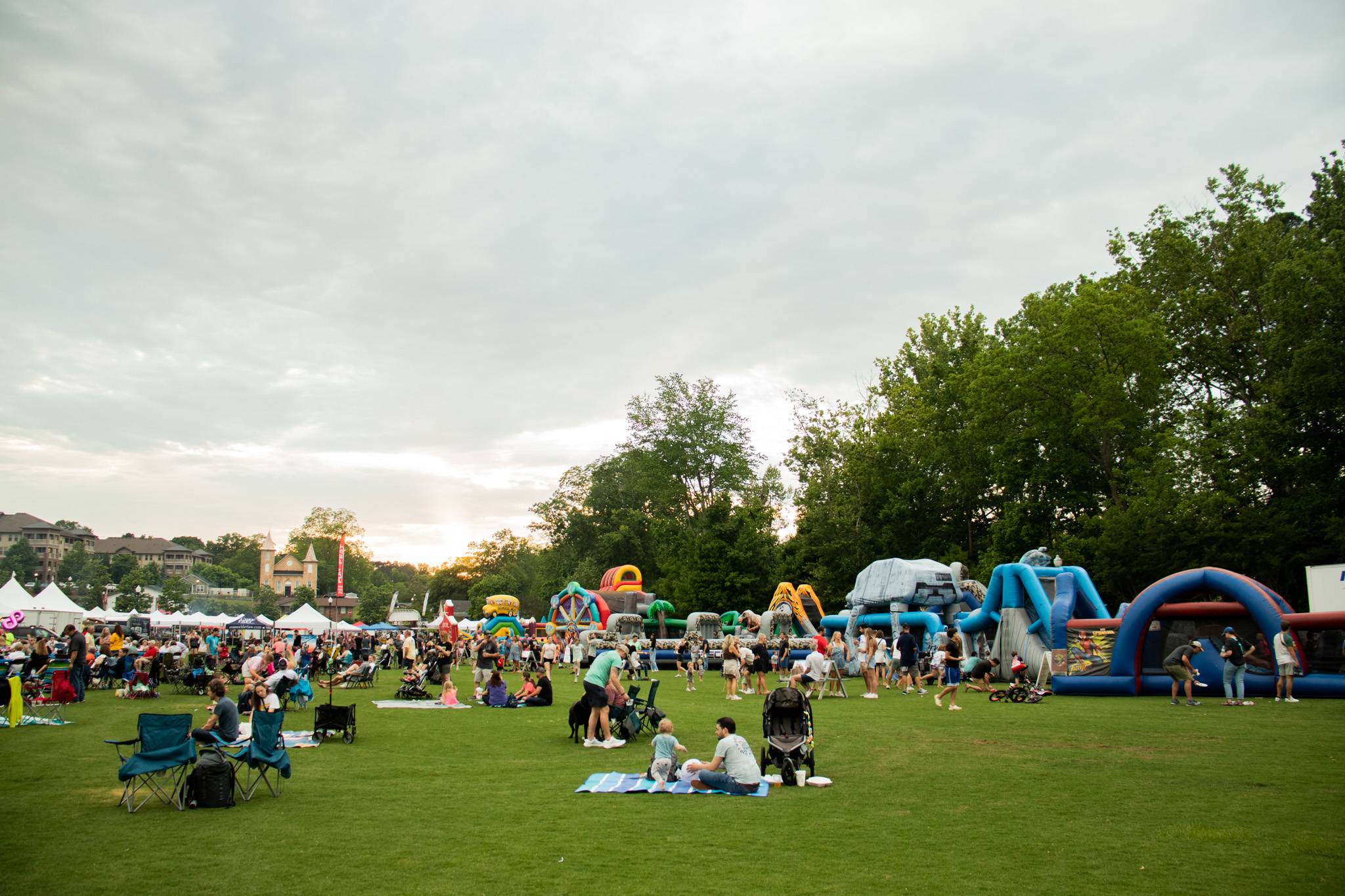 People gather on a grassy field with blankets and chairs at an outdoor event featuring tents and inflatable obstacle courses under a cloudy sky.