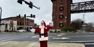 A person dressed as Santa Claus stands at an intersection in a small town, with decorated buildings and holiday wreaths visible in the background—your guide to Acworth gifts and festive cheer.