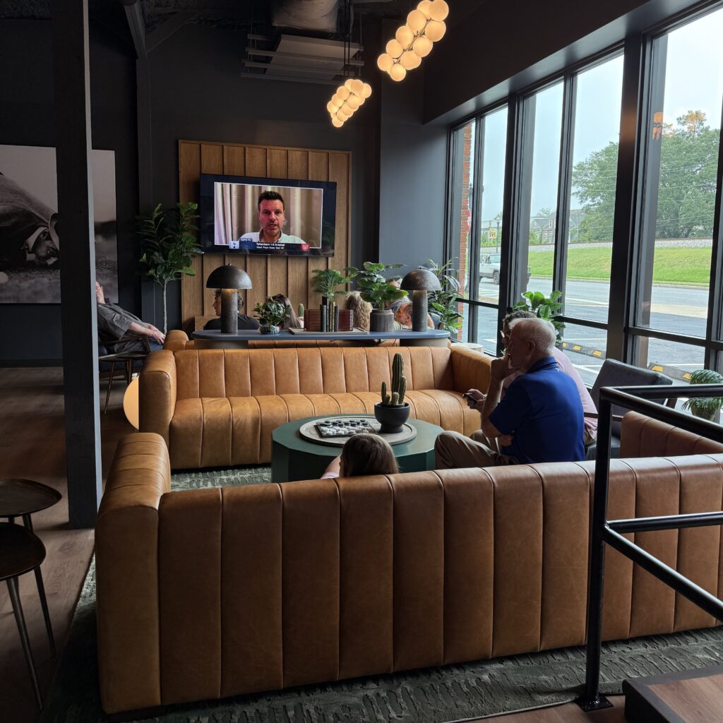 Three people sit on tan couches in a modern lounge, watching a man speak on a wall-mounted TV; large windows let in natural light, and plants decorate the room.