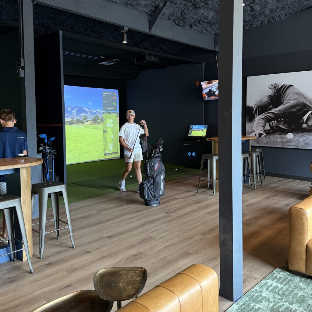 A person stands near a golf bag in an indoor golf simulator room with high tables, stools, a TV, and a large photo of a golfer on the wall.