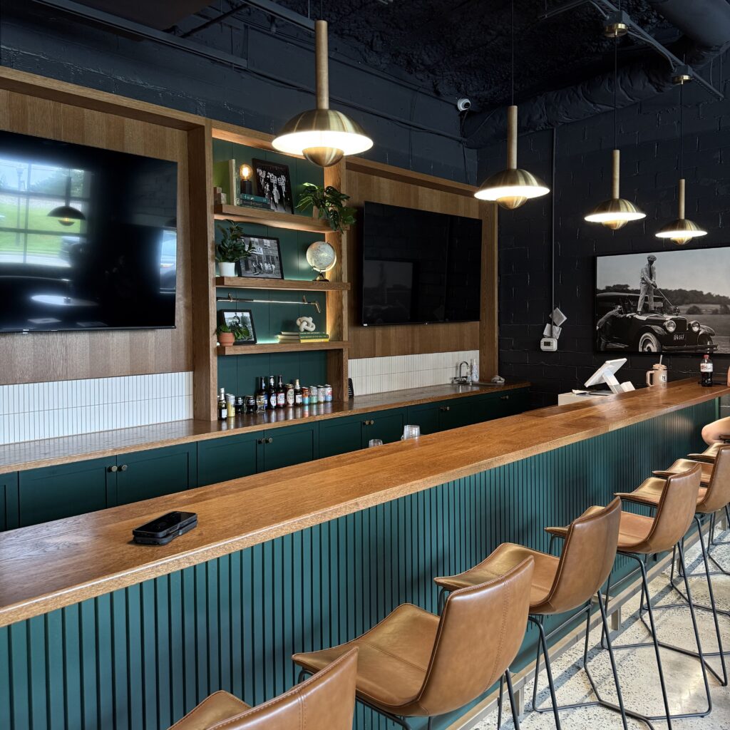 A modern bar with a long wooden counter, green paneling, leather chairs, hanging lights, shelves with decor, and two wall-mounted TVs.