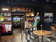 Photo Aug 01 2025, 11 14 31 AM Three people are talking near a counter inside a modern cafe with merch shelves, t-shirts on display, and high tables with stools.