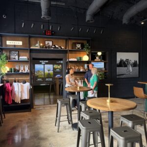 Three people stand and talk inside a modern store with merch shelves, t-shirts, hats, and stools around round wooden tables—your guide to Acworth gifts awaits among the unique finds.