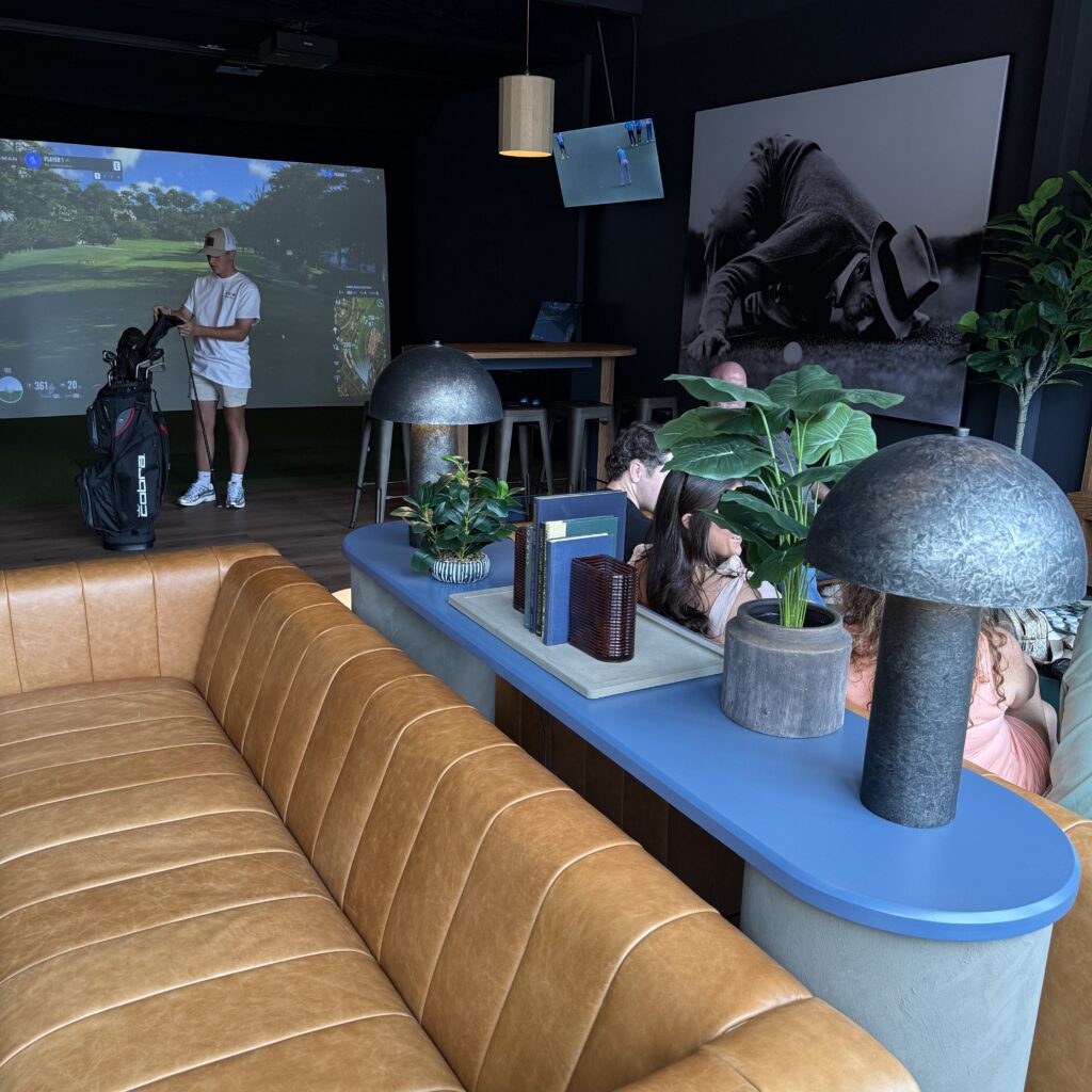 A man prepares golf clubs near a golf simulator screen while several people sit on a brown leather couch and blue table in a modern lounge with plants and decor.