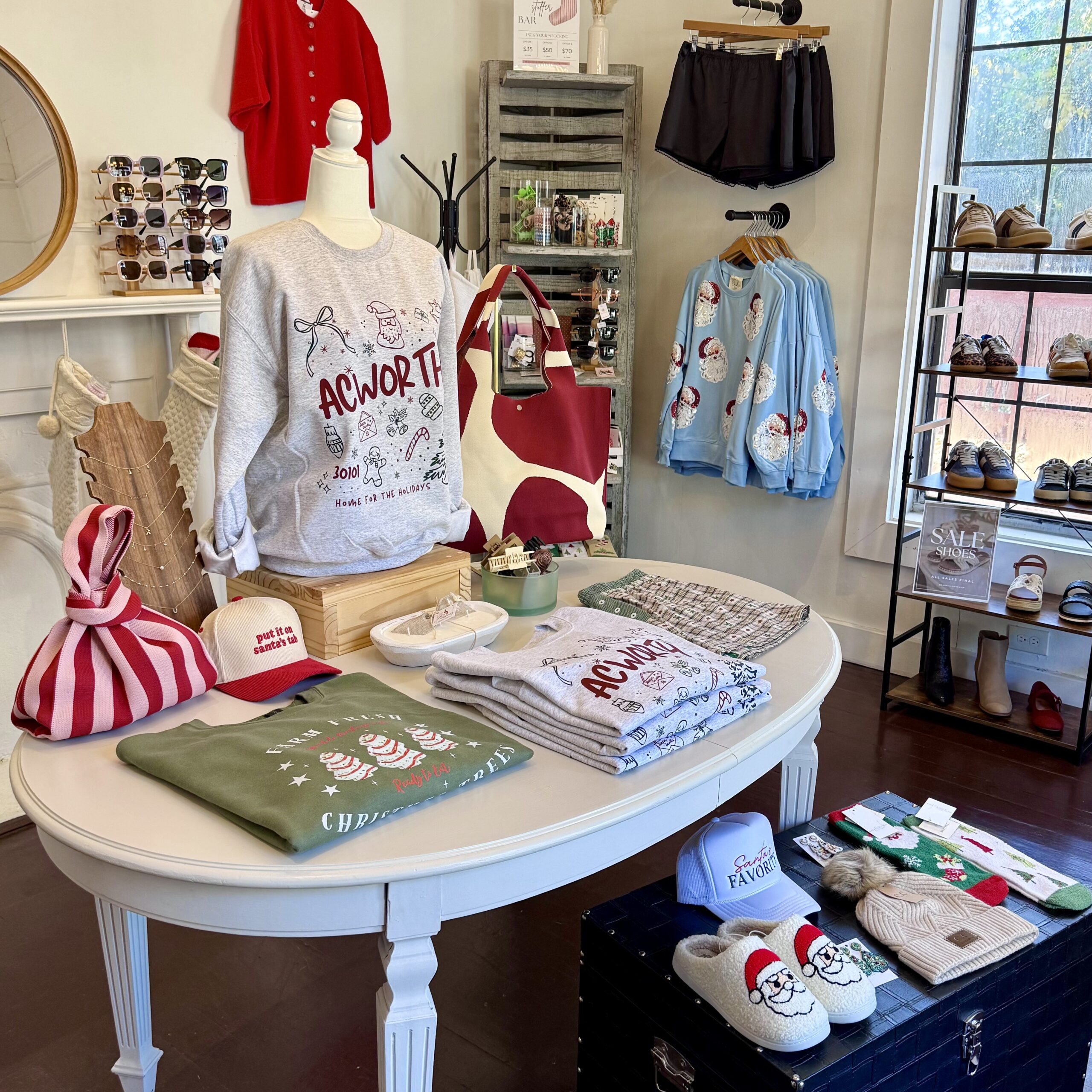 Clothing and accessories with holiday designs are displayed on tables and racks in a boutique, including sweaters, hats, pajamas, bags, and shoes—your guide to Acworth gifts this season.