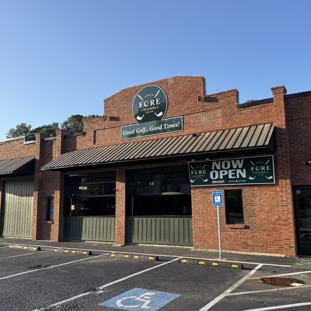 Exterior of a brick building with a “FORE Atlanta” sign, a “NOW OPEN” banner, and a handicapped parking space in front under a clear sky.