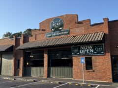 Photo Oct 02 2025, 8 32 12 AM Exterior of a brick building with a “FORE Atlanta” sign, a “NOW OPEN” banner, and a handicapped parking space in front under a clear sky.