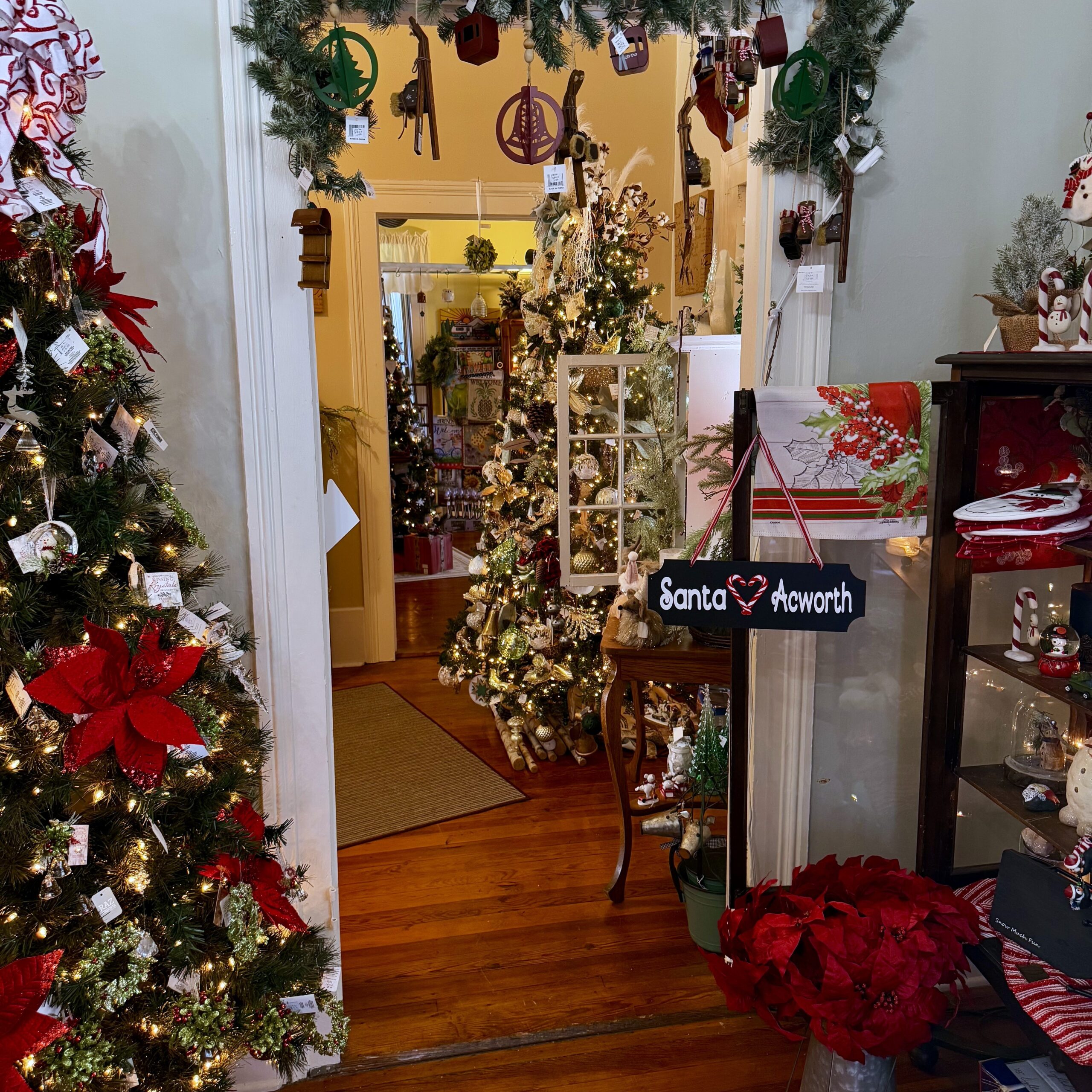 Room decorated for Christmas with multiple decorated trees, garlands, ornaments, poinsettias, and a sign reading "Santa ♥ Acworth"—your guide to Acworth gifts begins in this festive setting near the doorway.