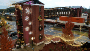 A brick building decorated with wreaths and snowflake lights stands near a railroad bridge as a freight train passes; golden holiday lights glow, creating the perfect scene for your guide to Acworth gifts.