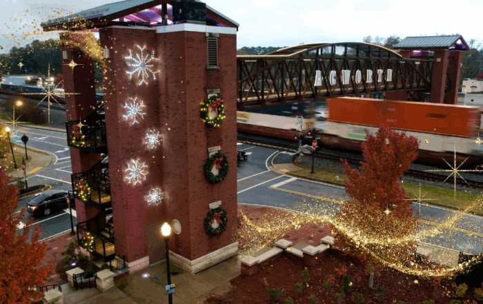 A brick building decorated with wreaths and snowflake lights stands near a railroad bridge as a freight train passes; golden holiday lights glow, creating the perfect scene for your guide to Acworth gifts.