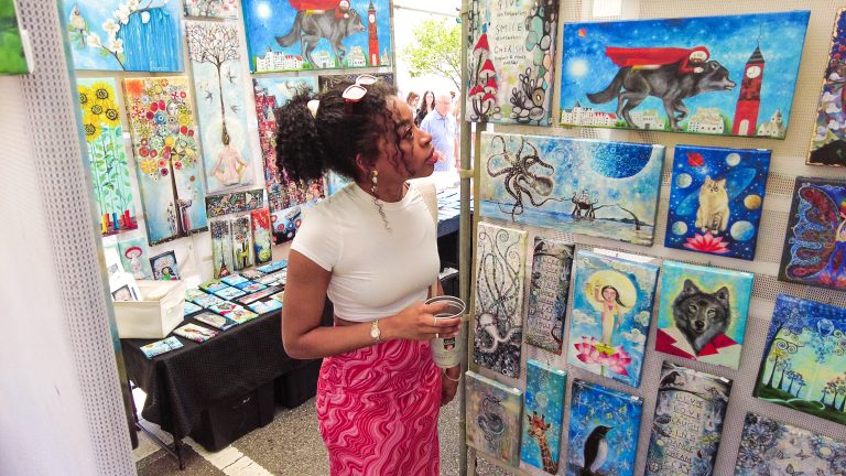 A woman with curly hair in a white top and pink skirt looks at colorful paintings displayed at an outdoor art market.