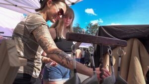 Two women at an outdoor market examine metal pans hanging from a wooden rack under a white canopy on a sunny day.