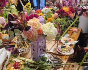 A worktable with assorted flowers, foliage, and tools for flower arranging; a floral arrangement in progress is in a vase at the center.