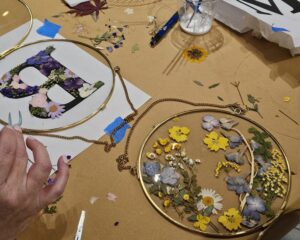 A hand arranges dried flowers on a circular frame, with pressed flowers, leaves, crafting tools, and a floral letter B on paper visible on the table.
