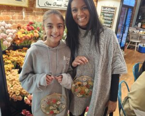 Two people standing indoors, smiling and holding circular frames with pressed flowers. They are surrounded by bouquets of flowers and a brick wall background.