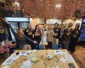 Nine women stand and pose behind a table with craft supplies, holding up circular flower art pieces inside a floral shop with a brick wall and flower arrangements.