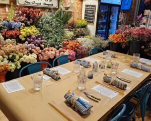 A table set with wrapped utensils, water glasses, and papers is arranged for a workshop in front of a large display of assorted flowers in a shop.